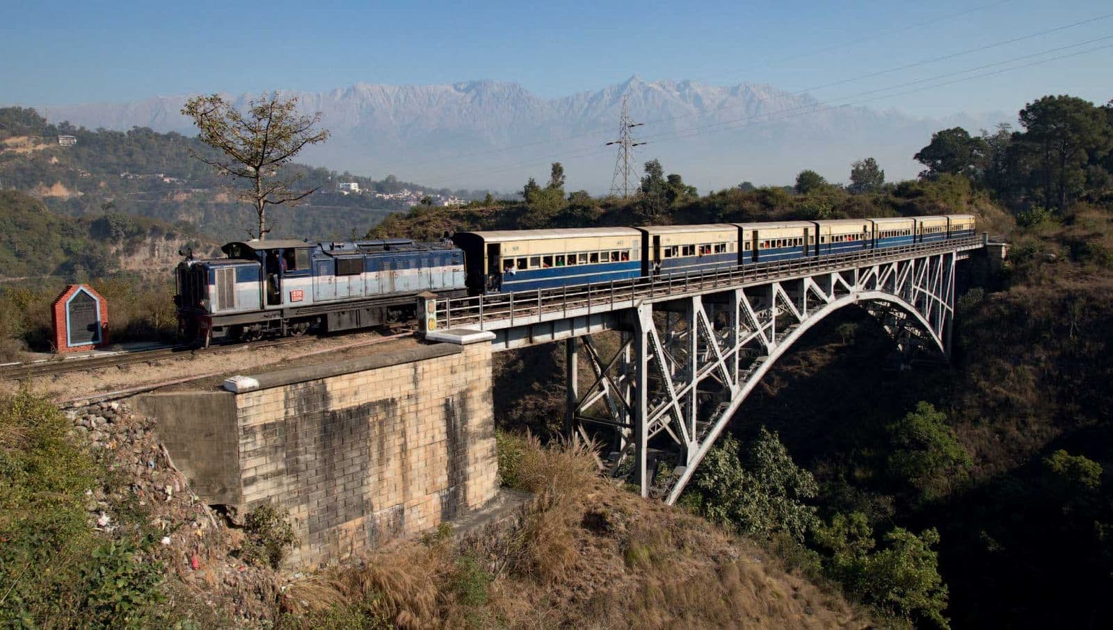 Kangra Valley Railway narrow-gauge train crossing the Reond Nullah arch bridge with the snow-capped Dhauladhar range behind
