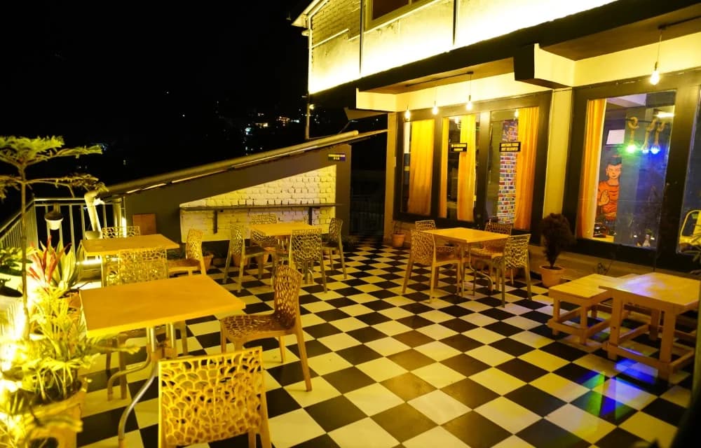 Terrace café at Friends by Alizeh at night, with yellow tables and chairs on a black-and-white chequered floor