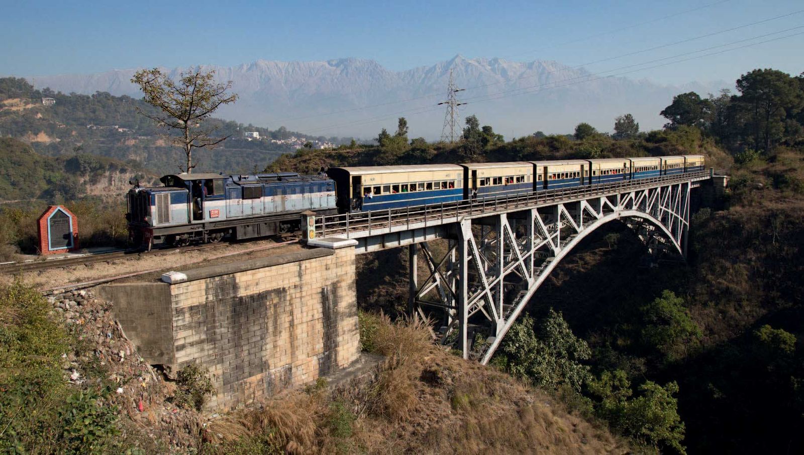 Kangra Valley Railway narrow-gauge train crossing the Reond Nullah arch bridge with the snow-capped Dhauladhar range behind