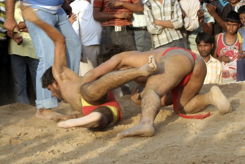 Two wrestlers grappling in a sand pit as spectators watch a traditional kushti bout in rural India